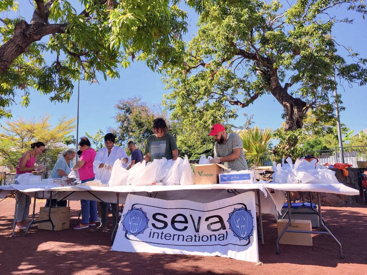 Des bénévoles préparant des sacs de repas sur une grande table, sous les arbres, devant une banderole SEVA International